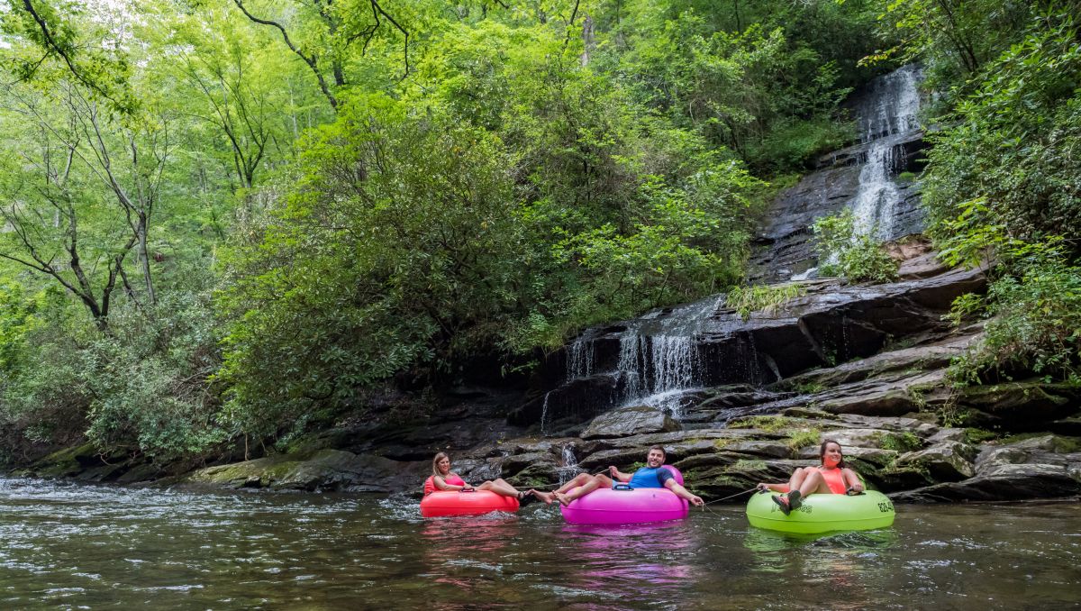 3 people floating down Deep Creek on colorful tubes with waterfall and green trees in background