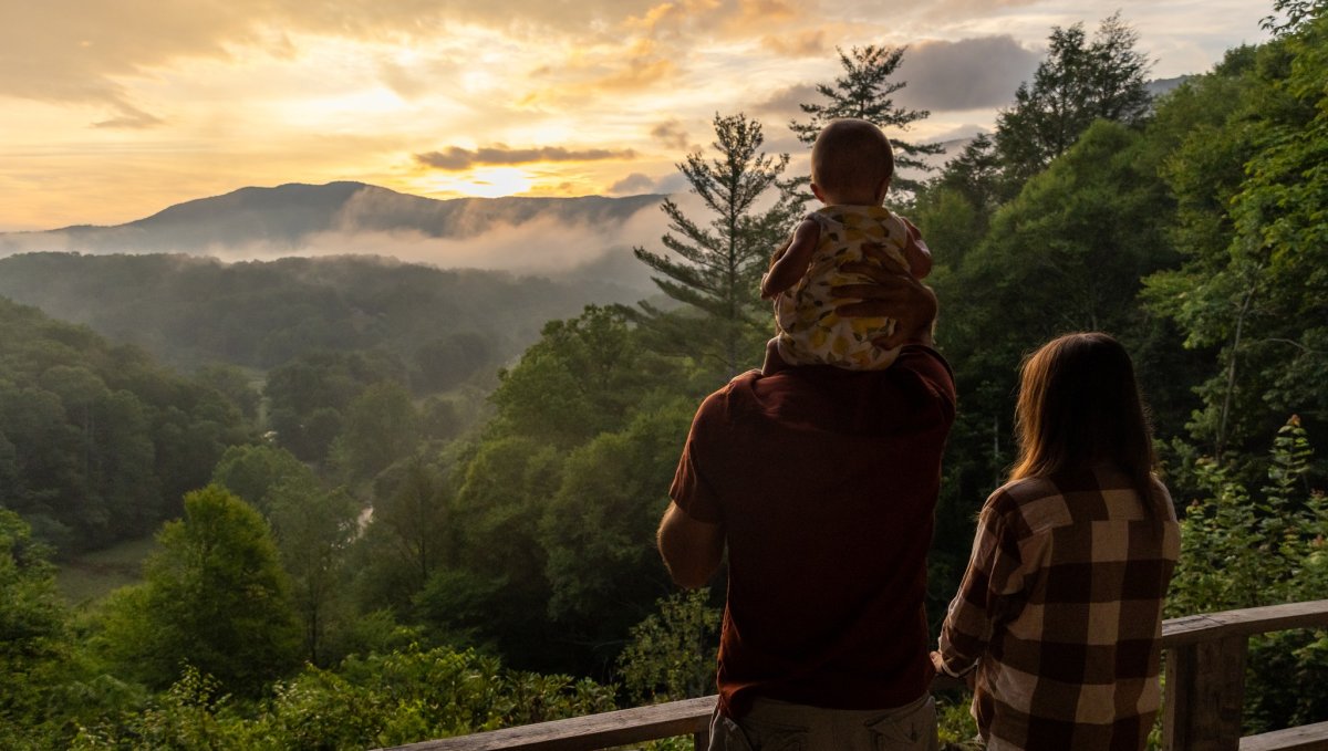 Couple with toddler on man's shoulders standing on porch overlooking mountains.