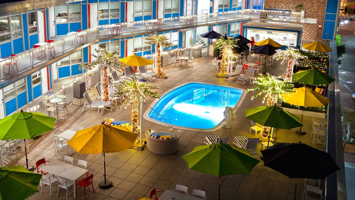 A vibrant retro-style hotel courtyard features a glowing pool surrounded by striped lounge chairs, colorful umbrellas, and twinkling lights wrapped around palm-like trees. The lively, modern atmosphere is illuminated against the evening sky.