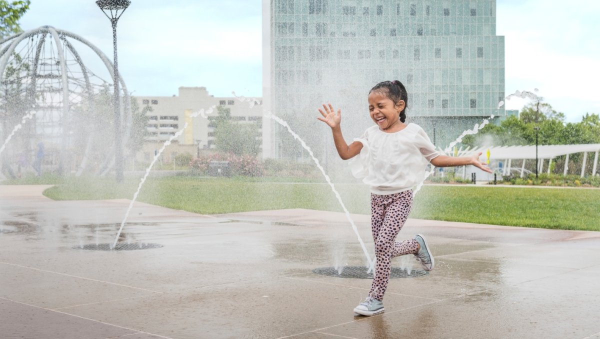 A young girl joyfully runs through a fountain with arcing streams of water on a sunny day, her laughter and excitement captured mid-motion. A modern building and urban park structures create a vibrant backdrop to the playful scene.