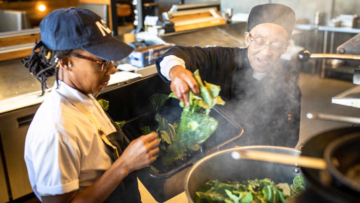 Two chefs placing collards into pot in restaurant's kitchen