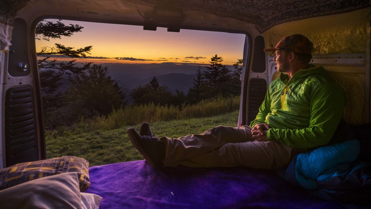 Man sitting in back of van and watching sunset over mountains in distance