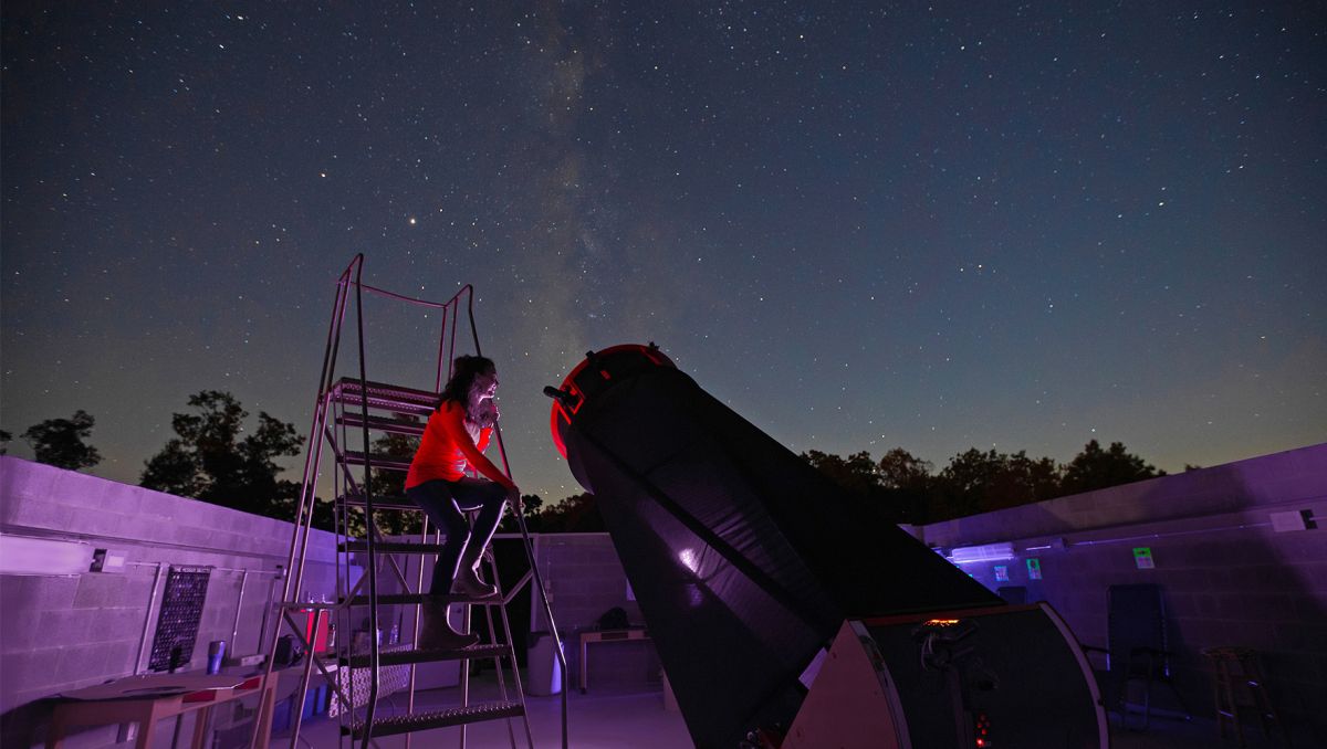 Person sitting on ladder and using telescope to look into starry night sky