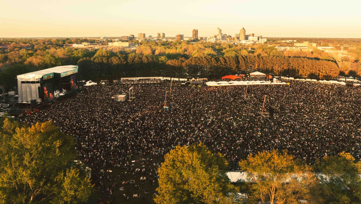 Aerial of large outdoor music festival crowd and stage with Raleigh skyline in background