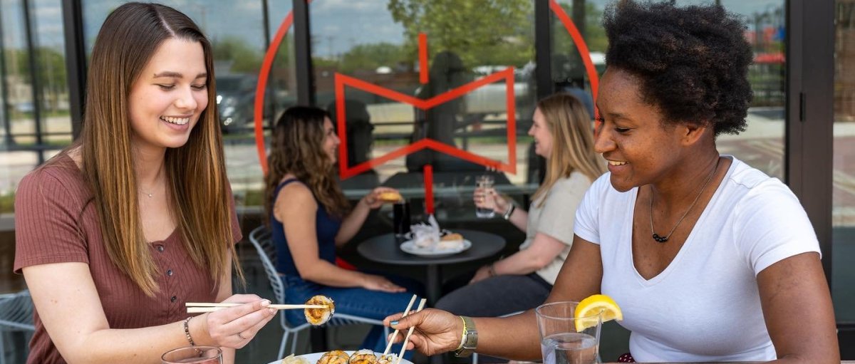 Two friends enjoying Asian food outdoors with more friends dining in background