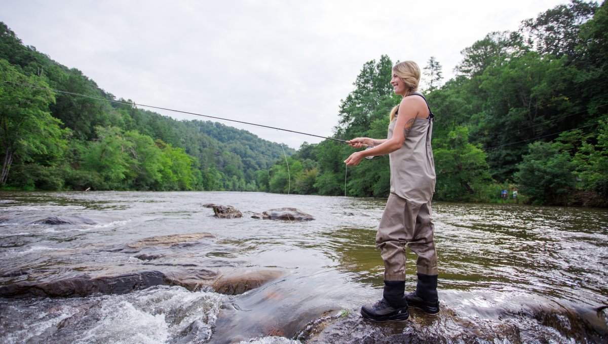 Woman fly-fishing in river surrounded by trees during daytime.