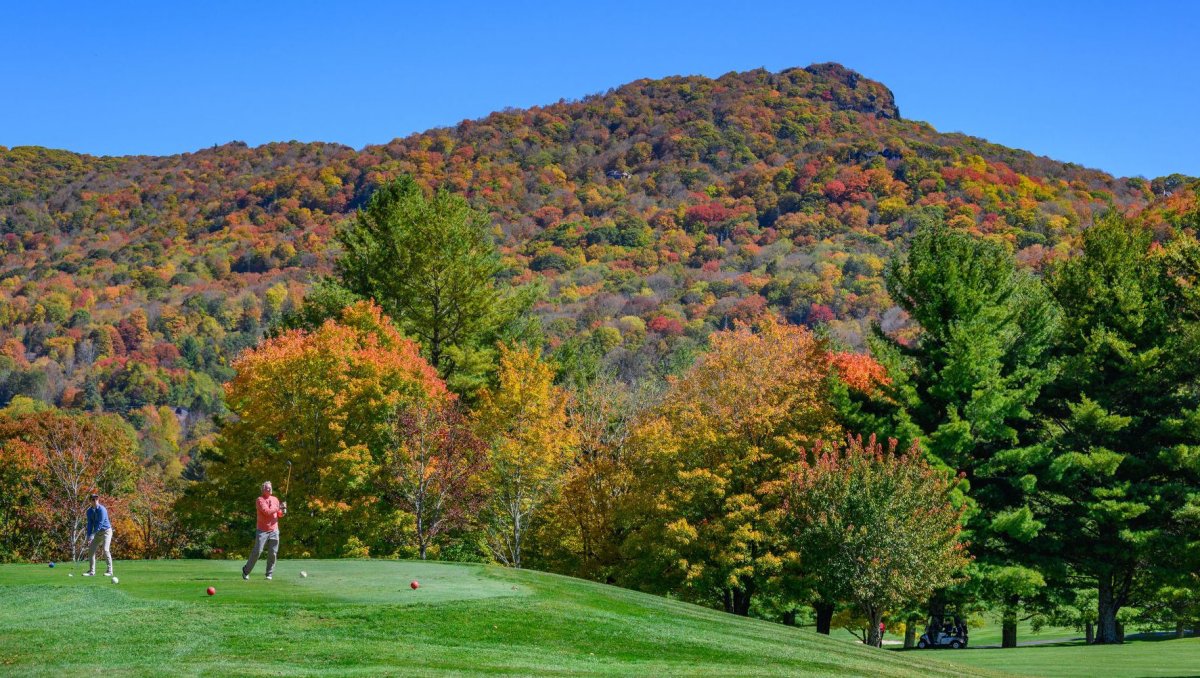 Two people teeing off on golf course with mountains with fall foliage in background