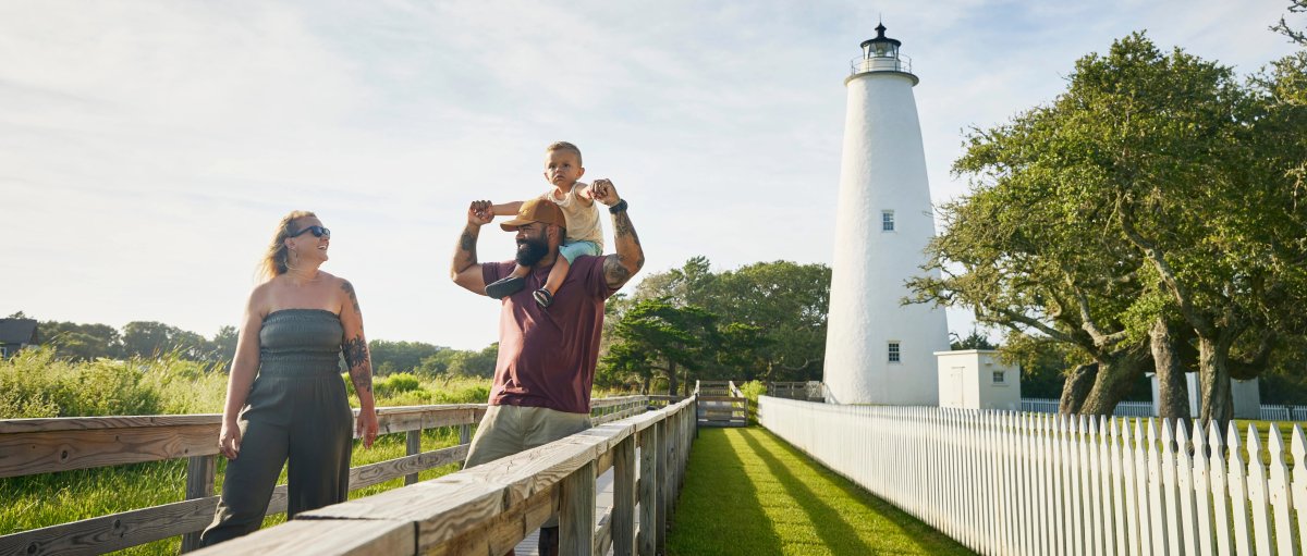 Family of three walking away from lighthouse, and father has kid on his shoulders, during daytime
