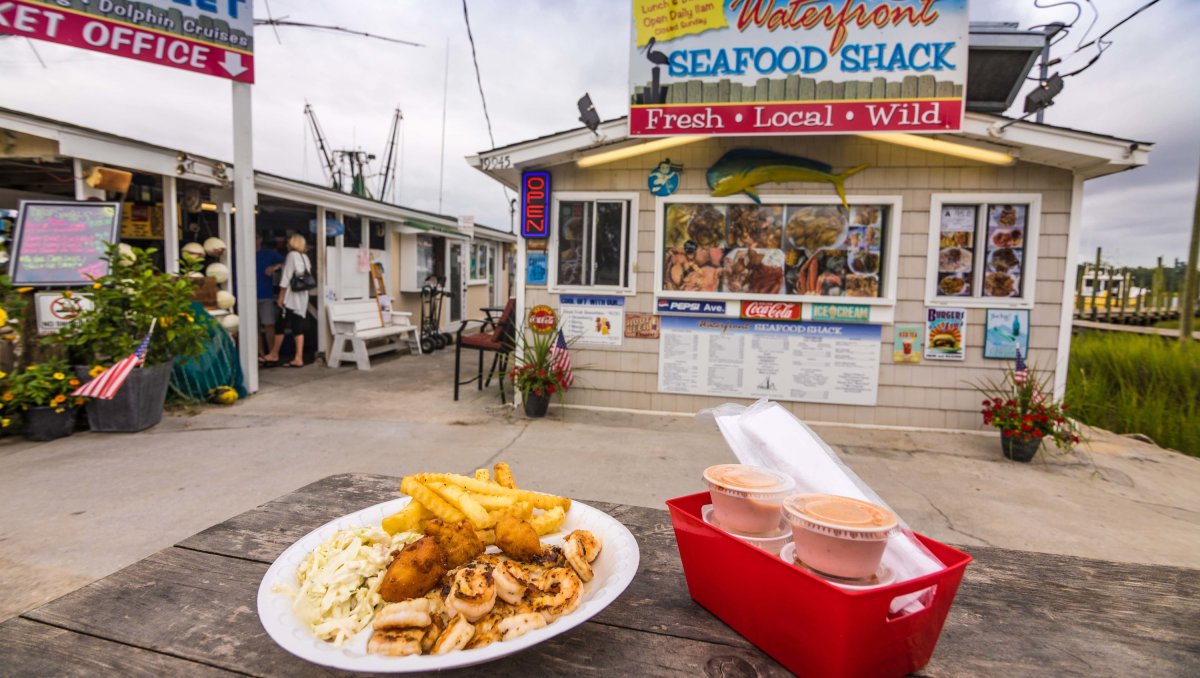 Plate of seafood plus sauces and utensils sitting on table in front of walk-up waterfront seafood shack