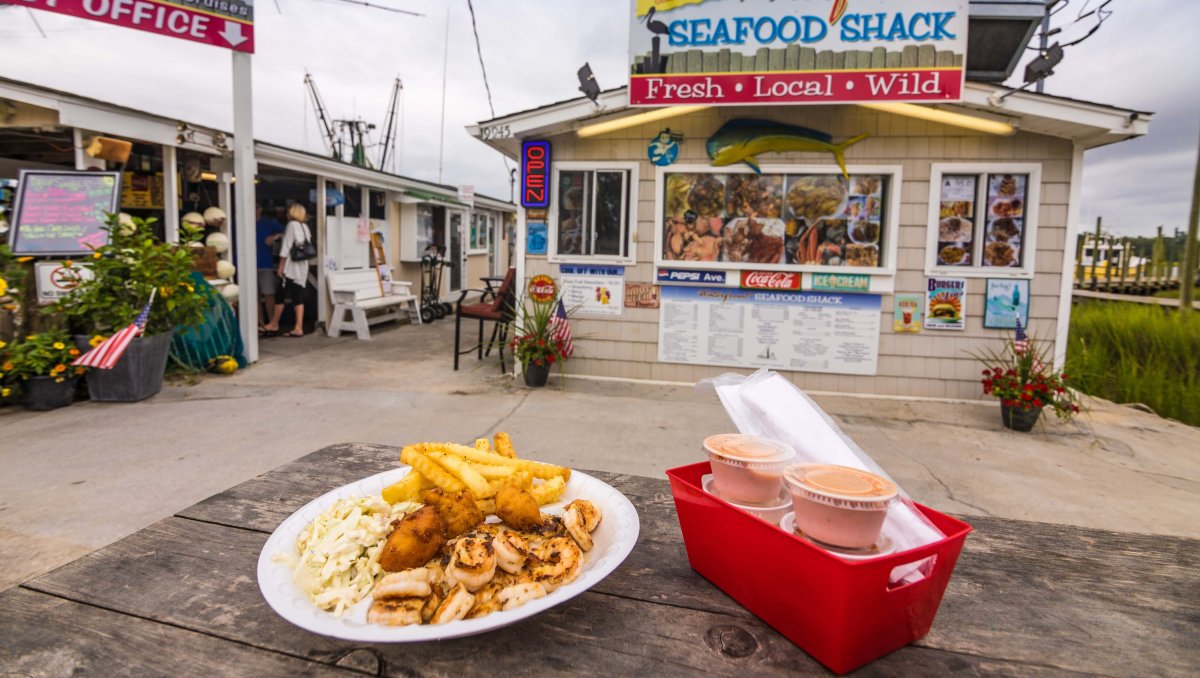 Plate of seafood plus sauces and utensils sitting on table in front of walk-up waterfront seafood shack