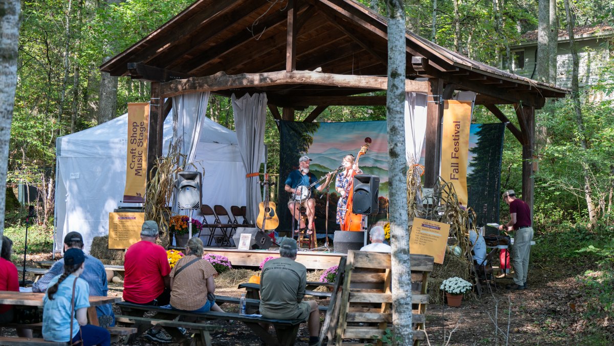 Band performing on outdoor stage at Folk School Fall Festival with forest in background and people sitting in seats in front.