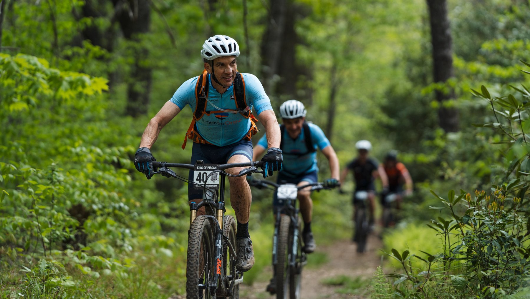 Four cyclers in a line on mountain trail surrounded by green trees