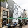 People sitting at tables under umbrellas and string lights with Earl Scruggs mural on side of building in background