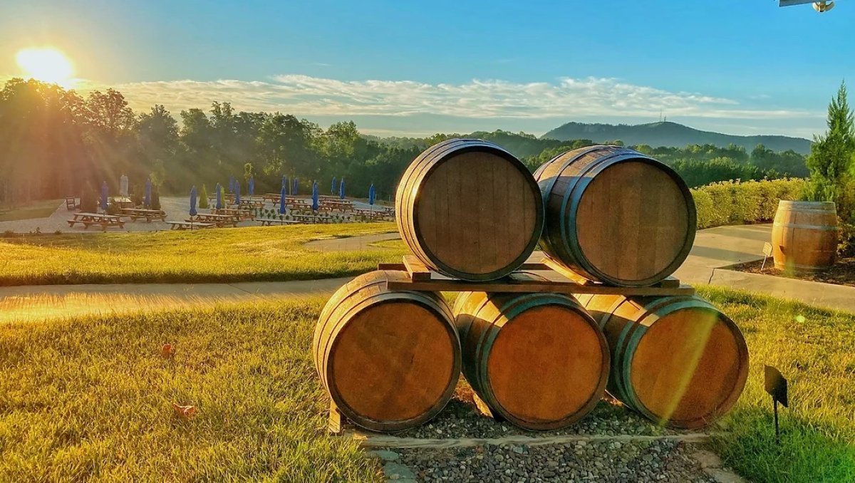 Wine barrels stacked in foreground with gorgeous winery grounds in background as sun sets behind trees.