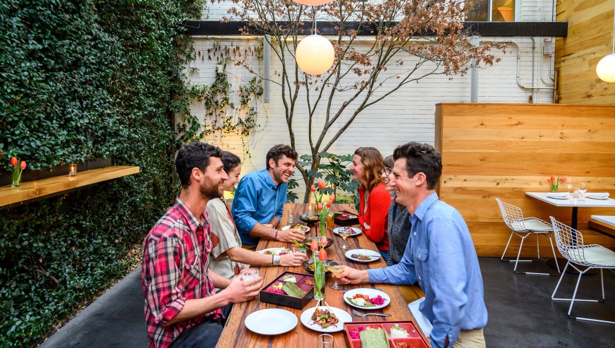 Friends sitting at wooden table outside enjoying meal surrounded by greenery wall and a tree.