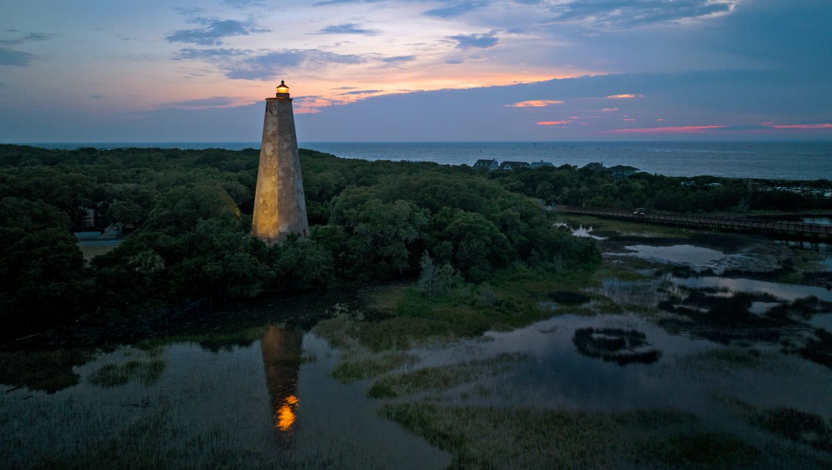 Old Baldy Lighthouse lit up at dusk surrounded by marsh and ocean.