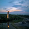 Old Baldy Lighthouse lit up at dusk surrounded by marsh and ocean.