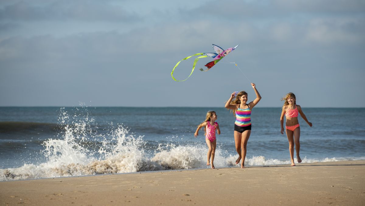 Three children running on the beach with the middle one flying a kit, waves crashing onto the sand behind them during daytime