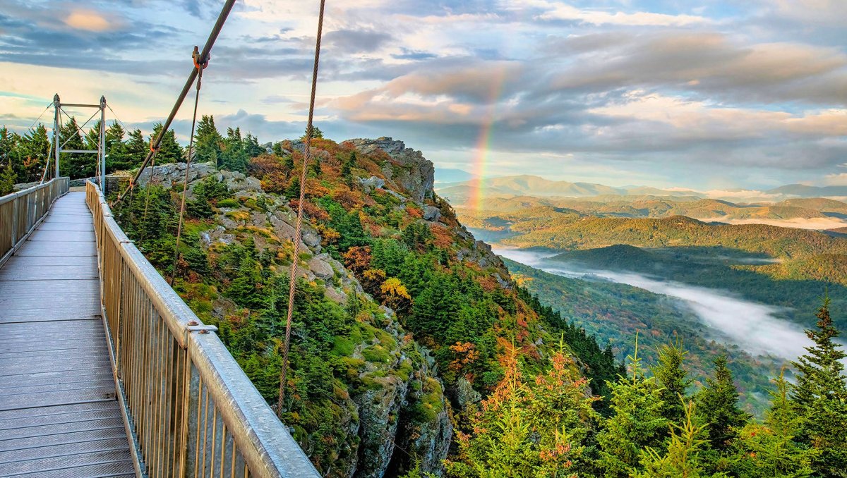 Suspension bridge extending to peak with fall foliage, mountains and a rainbow to the right