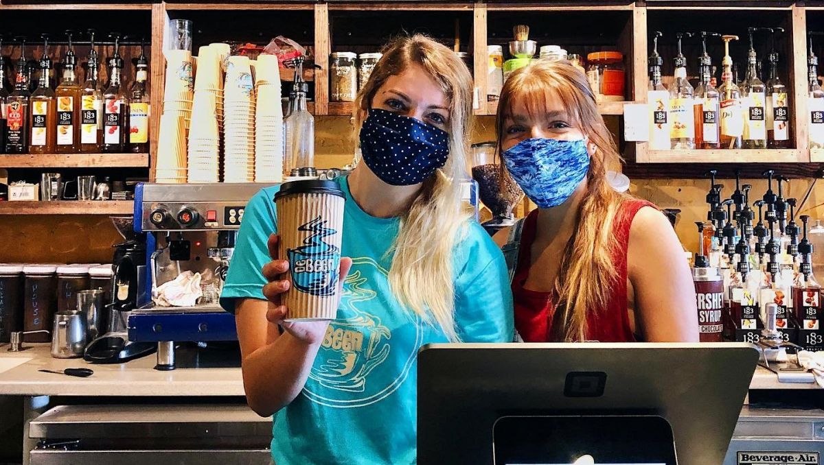 Two women wearing masks and holding up a coffee cup behind the Counter of DeBeen Espresso