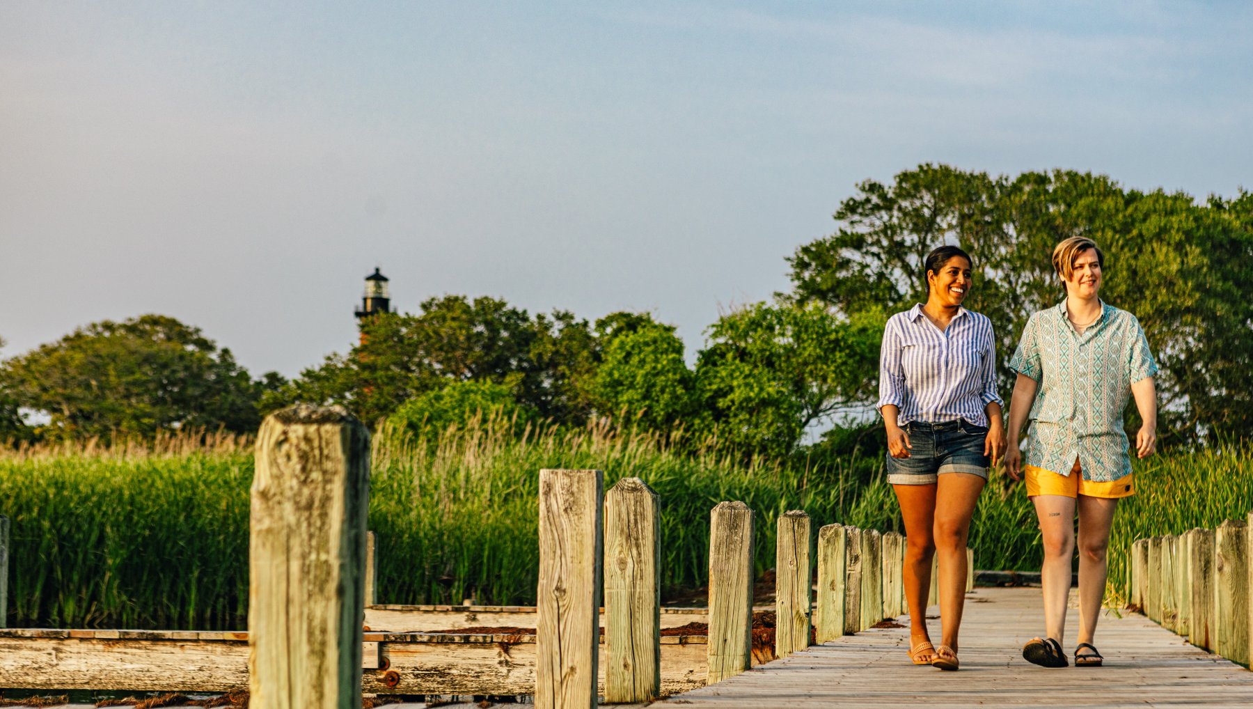 Couple wCouple walking down dock at Historic Corolla Park. alking on dock with green trees and lighthouse in background