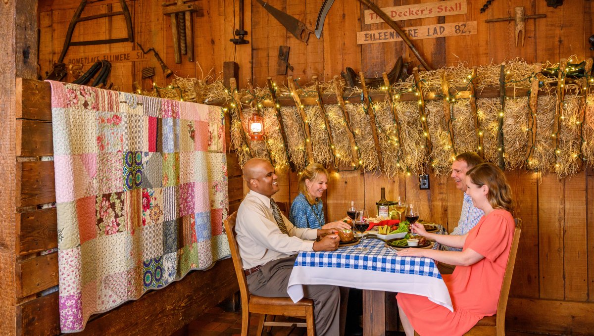 Group of diners at elegant barn restaurant surrounded by lights, hay and tools on wall.