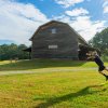 man mid-air tossing disc to basket in field in front of barn with sign: agricultural museum