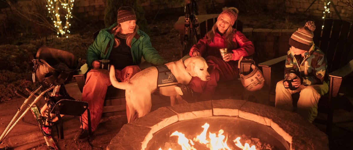 Family and dog sitting in front of outdoor fire pit with holiday lights in trees behind them