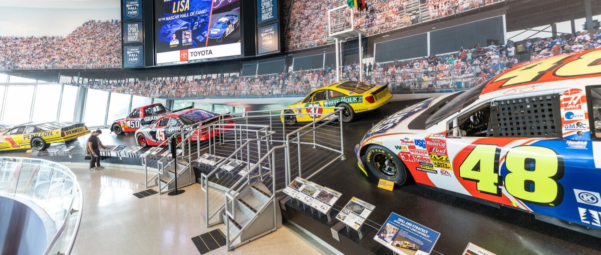 Museum exhibit of race cars and race trucks throughout the history of NASCAR, placed on a banking imitating a race track.