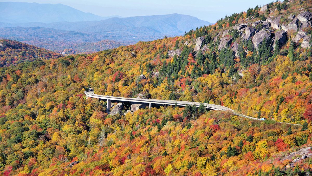 Aerial view of road winding through mountains surrounded by bright fall foliage and mountains in background