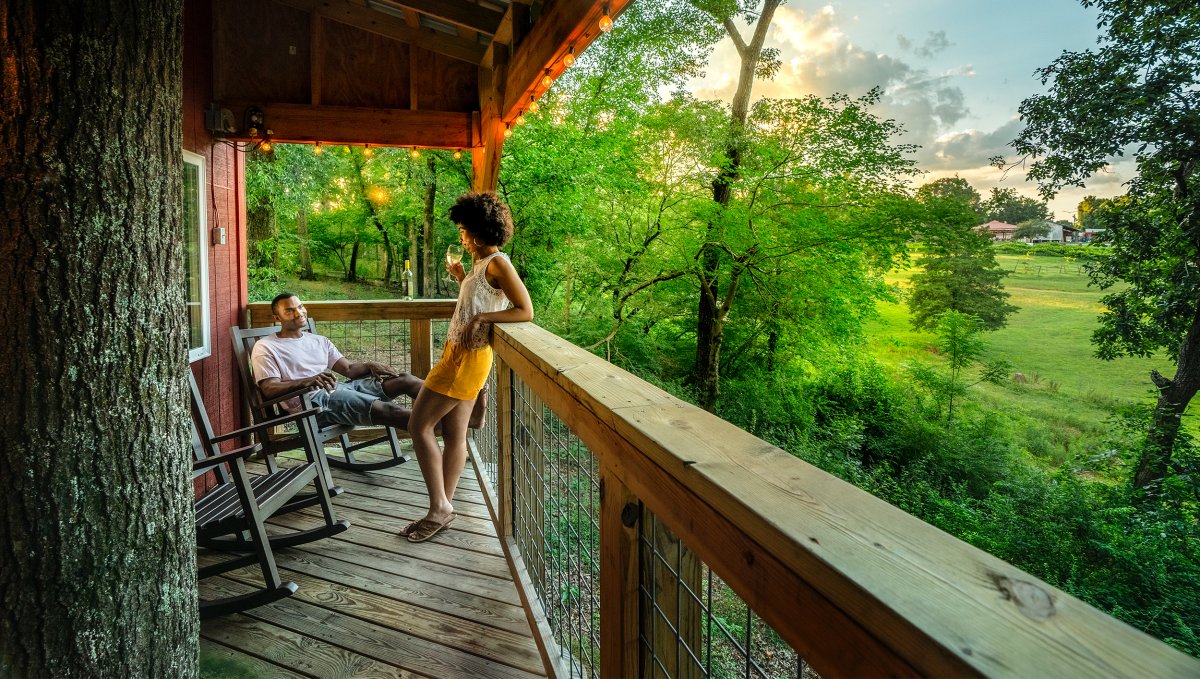 Man and woman drinking wine on the porch of a treehouse with green trees in background