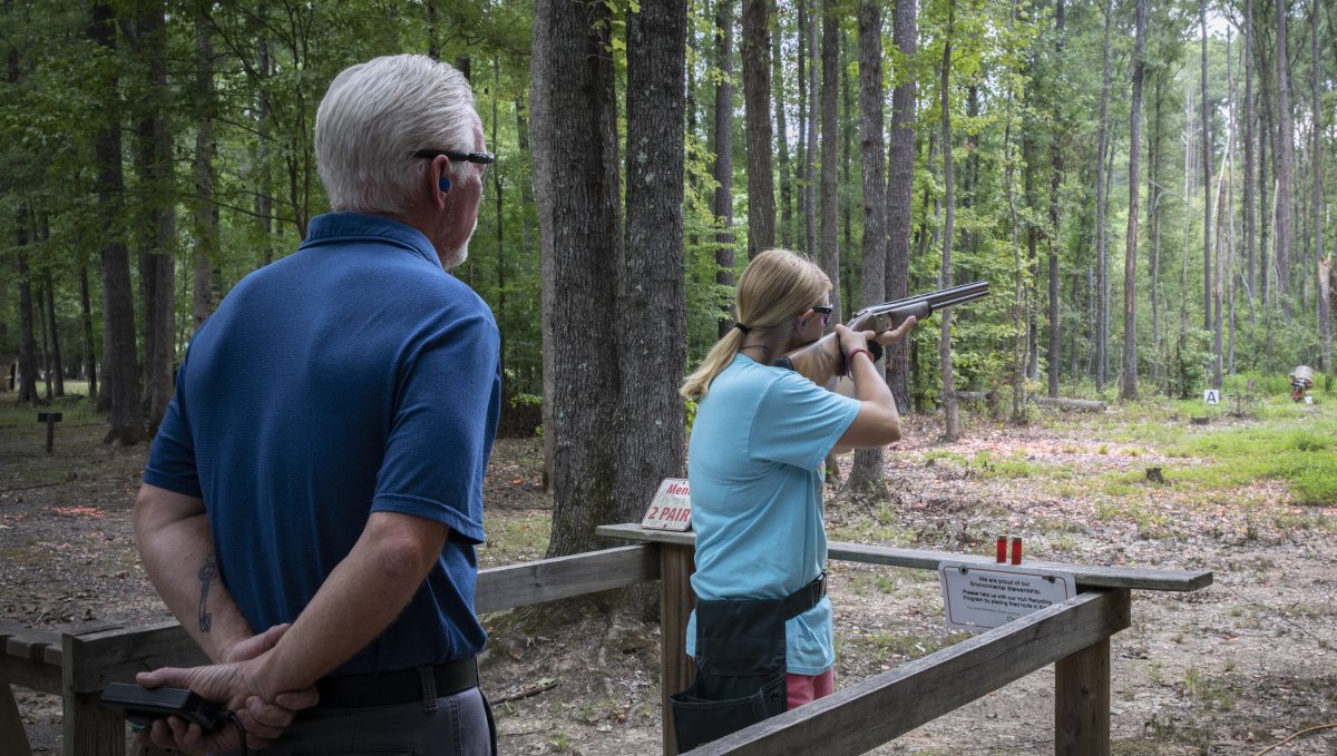 Woman shooting clays in woods as instructor watches on behind her.