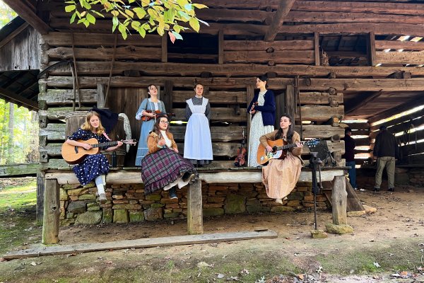 Women in period clothing singing and playing instruments in front of log cabin