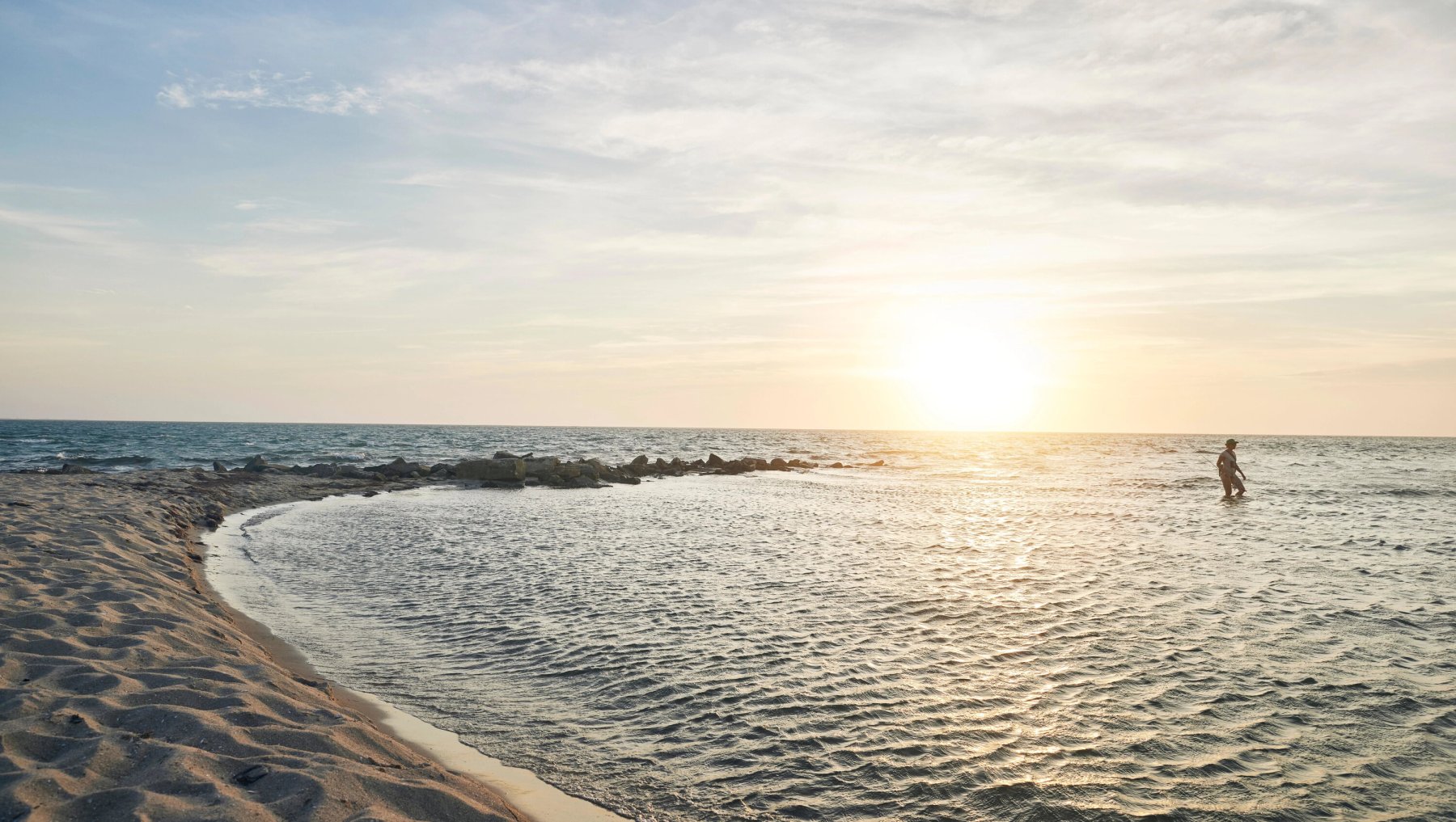 Person wading in water right off Springer's Point as sun sets in ocean in background.