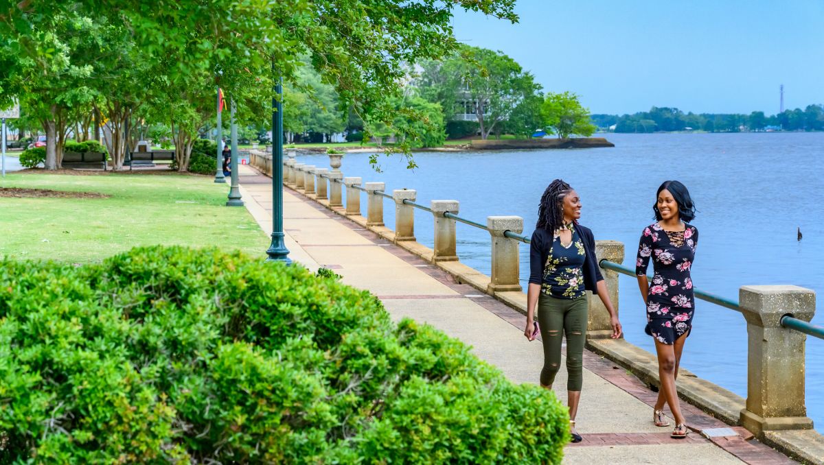Two women smiling at each other as they walk the New Bern waterfront with park to the left of them and water to the right
