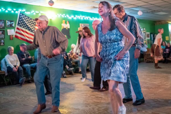 People line dancing in middle of dance floor with observers sitting along wall