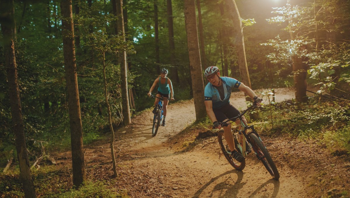 Two people cycling on Cub Creek Mountain Bike Trails with sun streaming through trees in forest