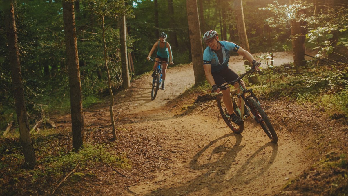 Two people cycling on Cub Creek Mountain Bike Trails with sun streaming through trees in forest