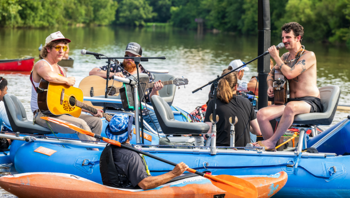 Band performing on river raft with people in kayaks surrounding them during on-water concert