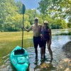 Couple standing in shallow river next to kayak, surrounded by green trees