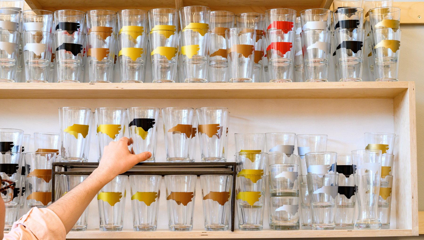 Close interior landscape of person's hand reaching for pint glass featuring North Carolina decals on display at DECO retail shop in downtown Raleigh.