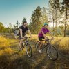 Smiling couple on bikes pedaling through grass trail during sunset