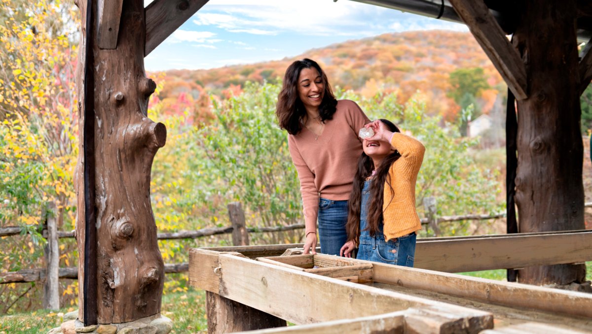 A smiling mother and daughter enjoy a gem mining activity under a rustic wooden shelter. The daughter holds a sparkling gem up to her eye, beaming with excitement, while autumn foliage and rolling hills create a colorful backdrop.