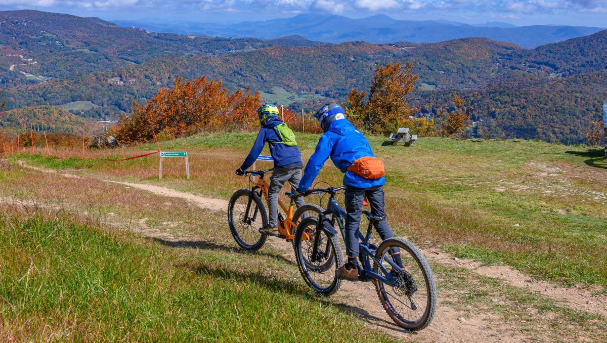 Two friends in blue jackets riding bikes on mountain with mountains with fall foliage in background