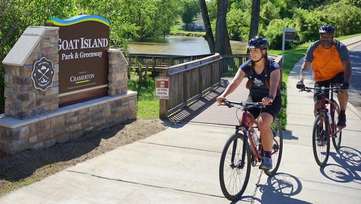 Two people biking on sidewalk in front of sign reading Goat Island with water and trees in background.