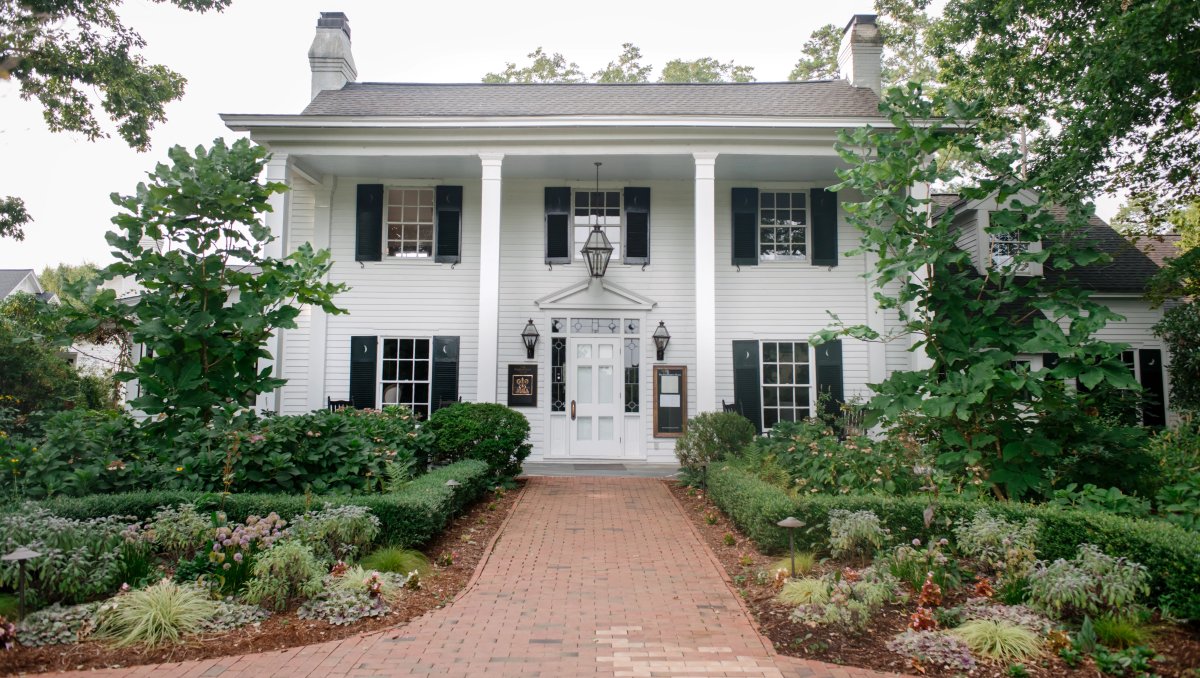 Front exterior of a beautiful white inn, with gardens and brick sidewalk in front during daytime