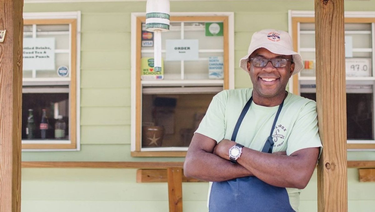 A man stands against a wooden pole and smiles. Three order windows sit behind him.