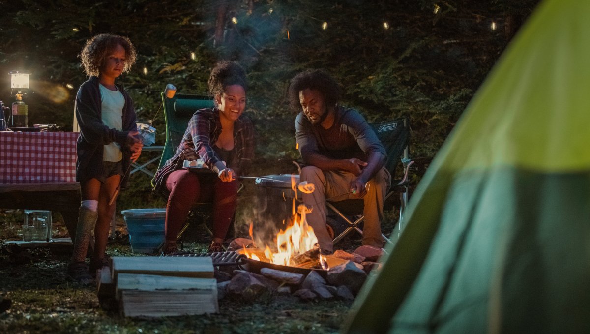 Family of three roasting marshmallows over campfire at night with tent in foreground.