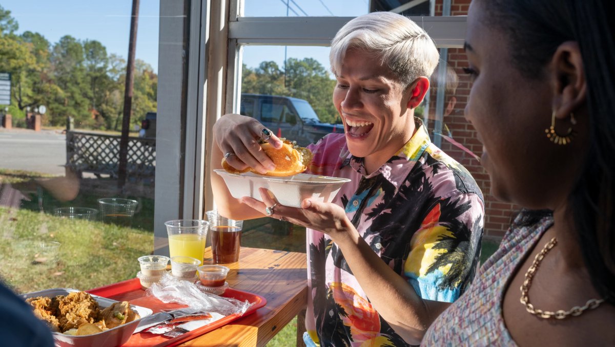 Person sitting at table about to bite into seafood sandwich with plate of food in front