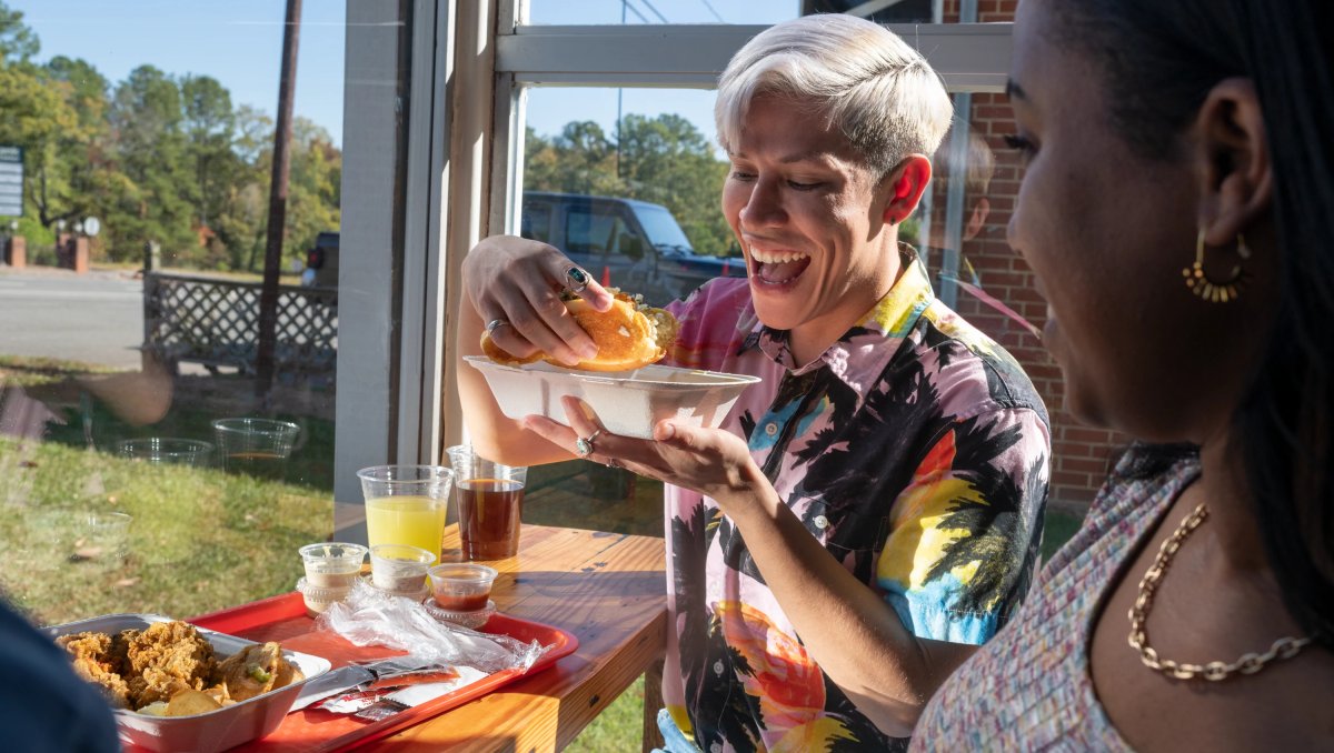 Person sitting at table about to bite into seafood sandwich with plate of food in front